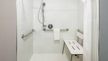 Un espace douche avec un sol carrelé de carreaux blancs, une barre d’appui, un porte-serviettes et une pomme de douche.