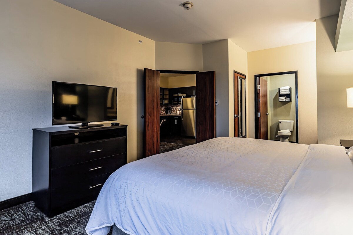 A hotel room with a bed, a TV, a dresser, a bathroom, and a kitchen area.