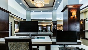Un hall de hotel con una recepción, dos estaciones de trabajo con computadora, un televisor de pantalla plana grande y un área de comedor con candelabro.