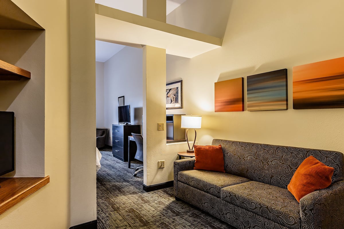 A hotel room with a TV on a wooden stand, a patterned sofa with orange cushions, a desk with a chair, and artwork on the walls.
