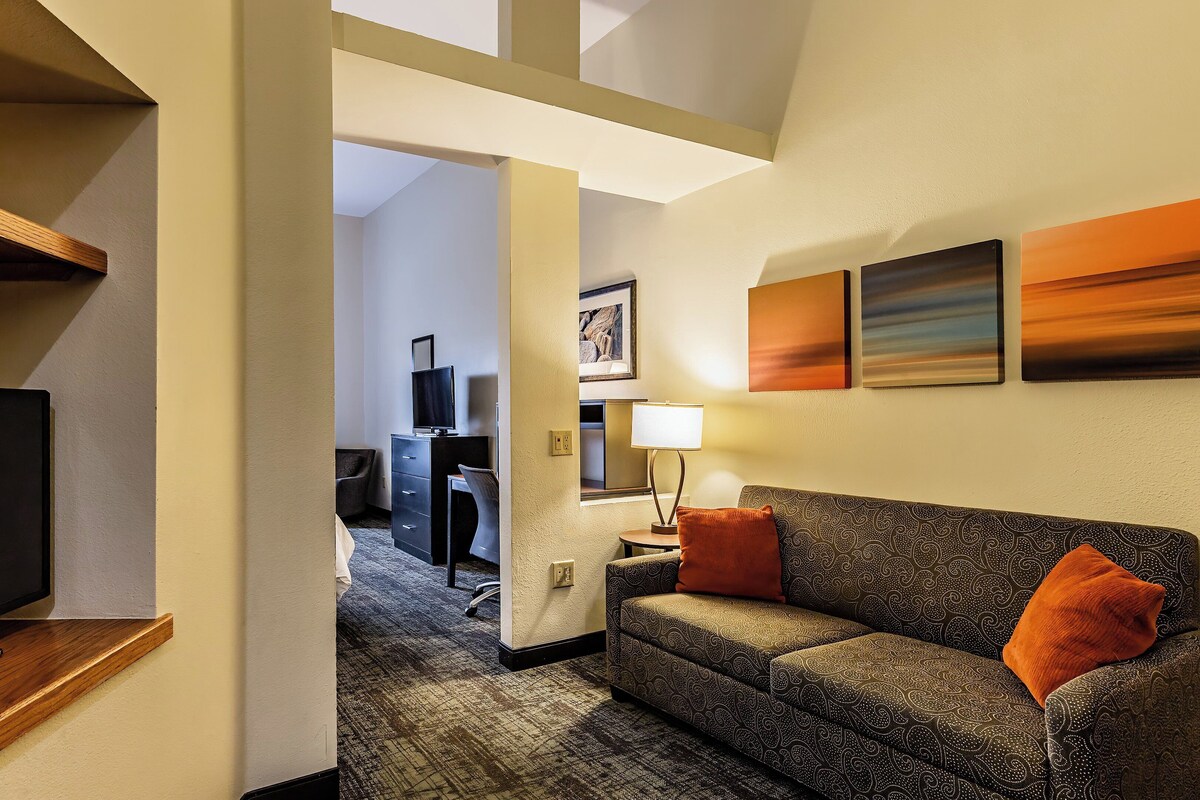 A hotel room with a TV on a wooden stand, a patterned sofa with orange cushions, a desk with a chair, and artwork on the walls.