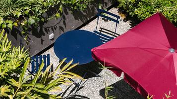 Un área de patio con una mesa y sillas azules, rodeada de exuberante follaje y un paraguas rojo.