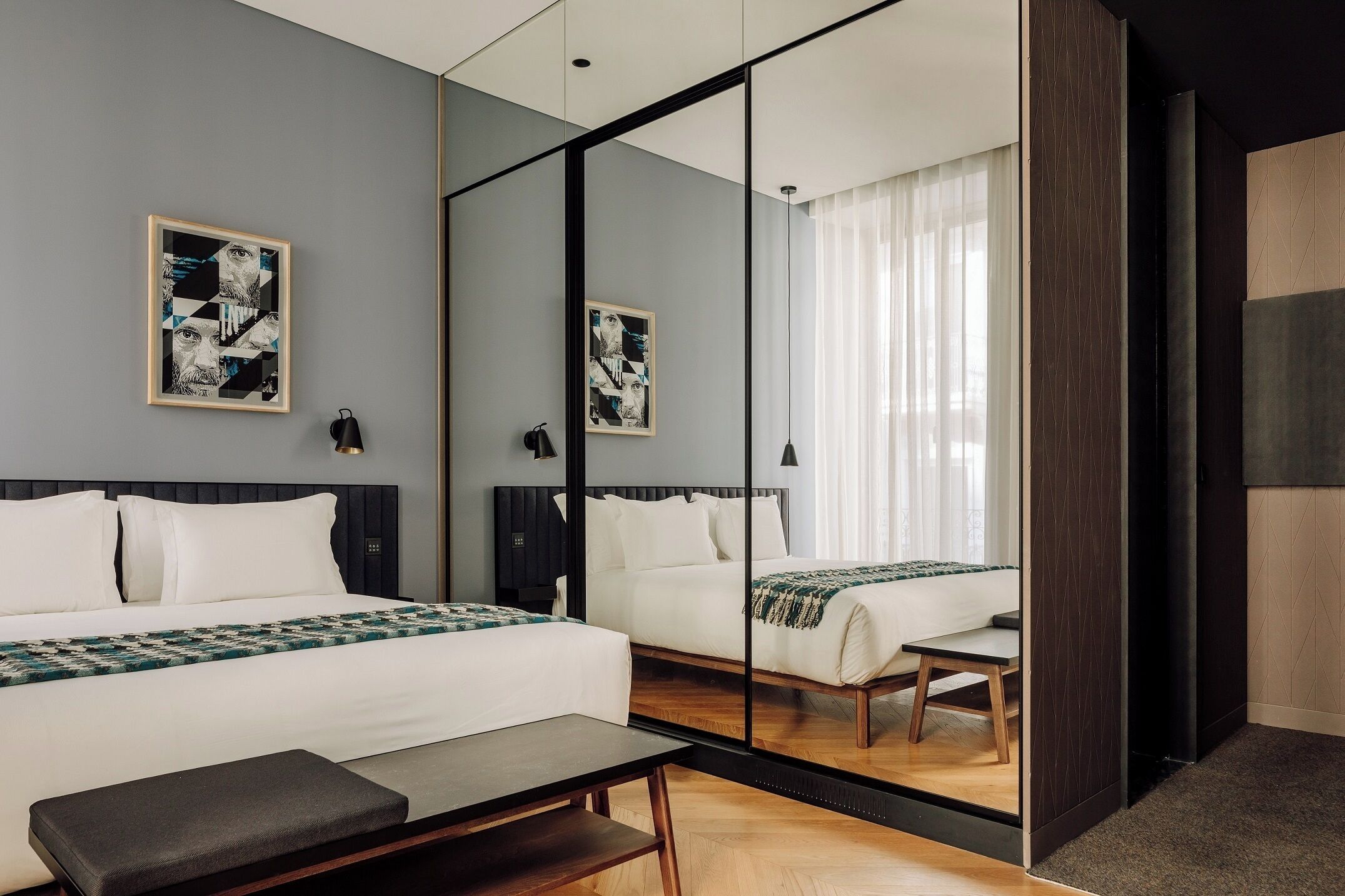 Una habitación de hotel moderna con un espejo grande que refleja la cama y un escritorio. Hay dos camas, un banco y una mesita.