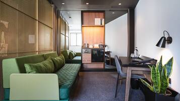 Ein modernes Hotelzimmer mit einem grünen Sofa, einem Schreibtisch mit einem Laptop, einer Zimmerpflanze und einem durch eine Schiebetür einsehbarer Küchenbereich.