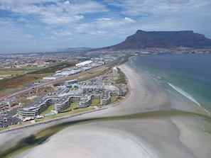 An aerial view of a coastal area with a prominent mountain, a beach, and a developed urban area with modern buildings and a road. - Sunstays Lagoon Beach (Cape Town)