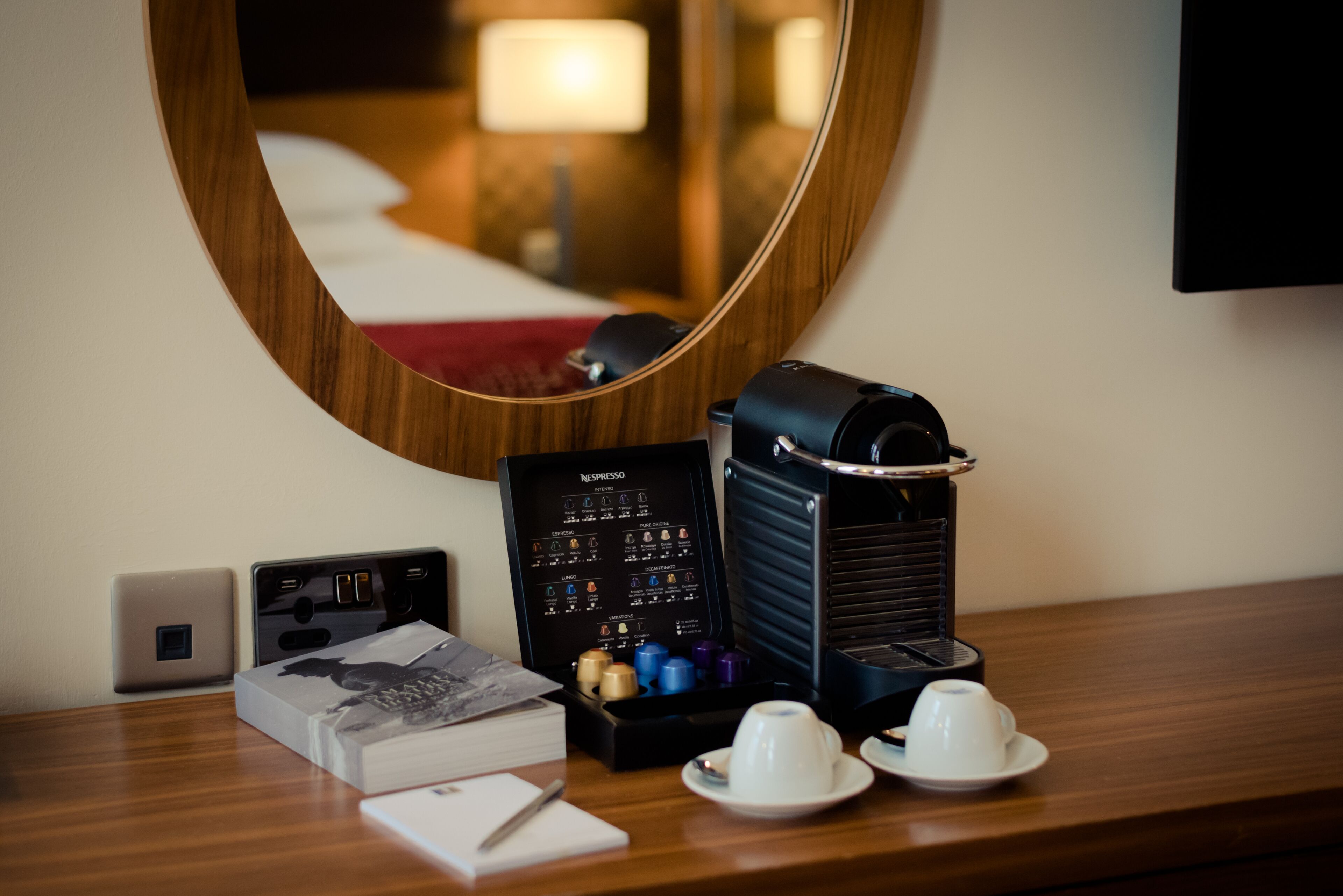 Hotelkamer met een houten bureau, een koffieapparaat, een display voor Nespresso-cups, een laptop en een boek.