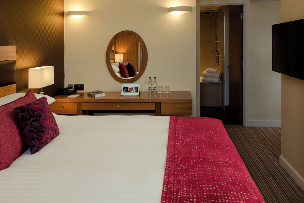 A hotel room with a large bed, a wooden dresser, a mirror, a bedside lamp, a TV, and a shower area.
