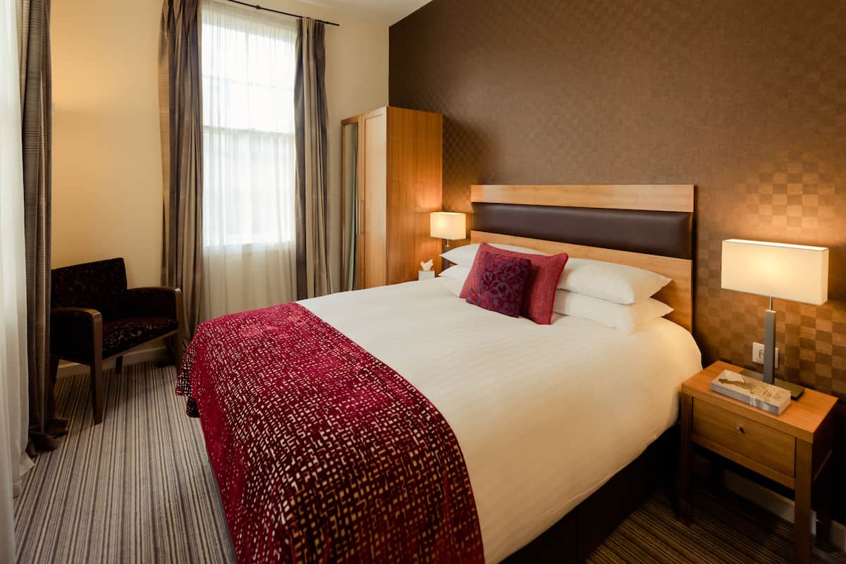 A hotel room with a large bed, a desk, a chair, and a wardrobe.