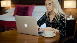 Une personne est assise à une table, un ordinateur portable devant elle, et une tasse de café à portée de main, dans une pièce où se trouvent un lit et des lampes.