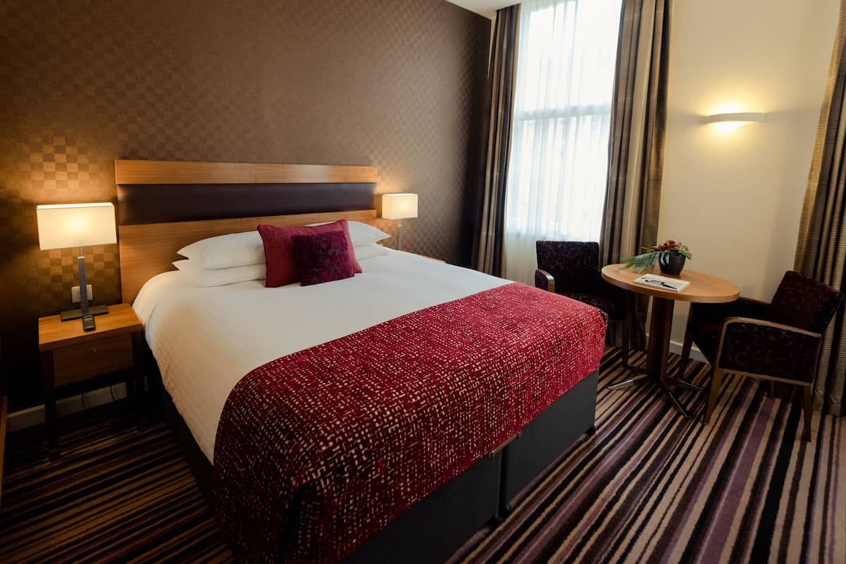 A hotel room with a large bed, two bedside tables with lamps, a small round table with a chair, and a window with curtains.