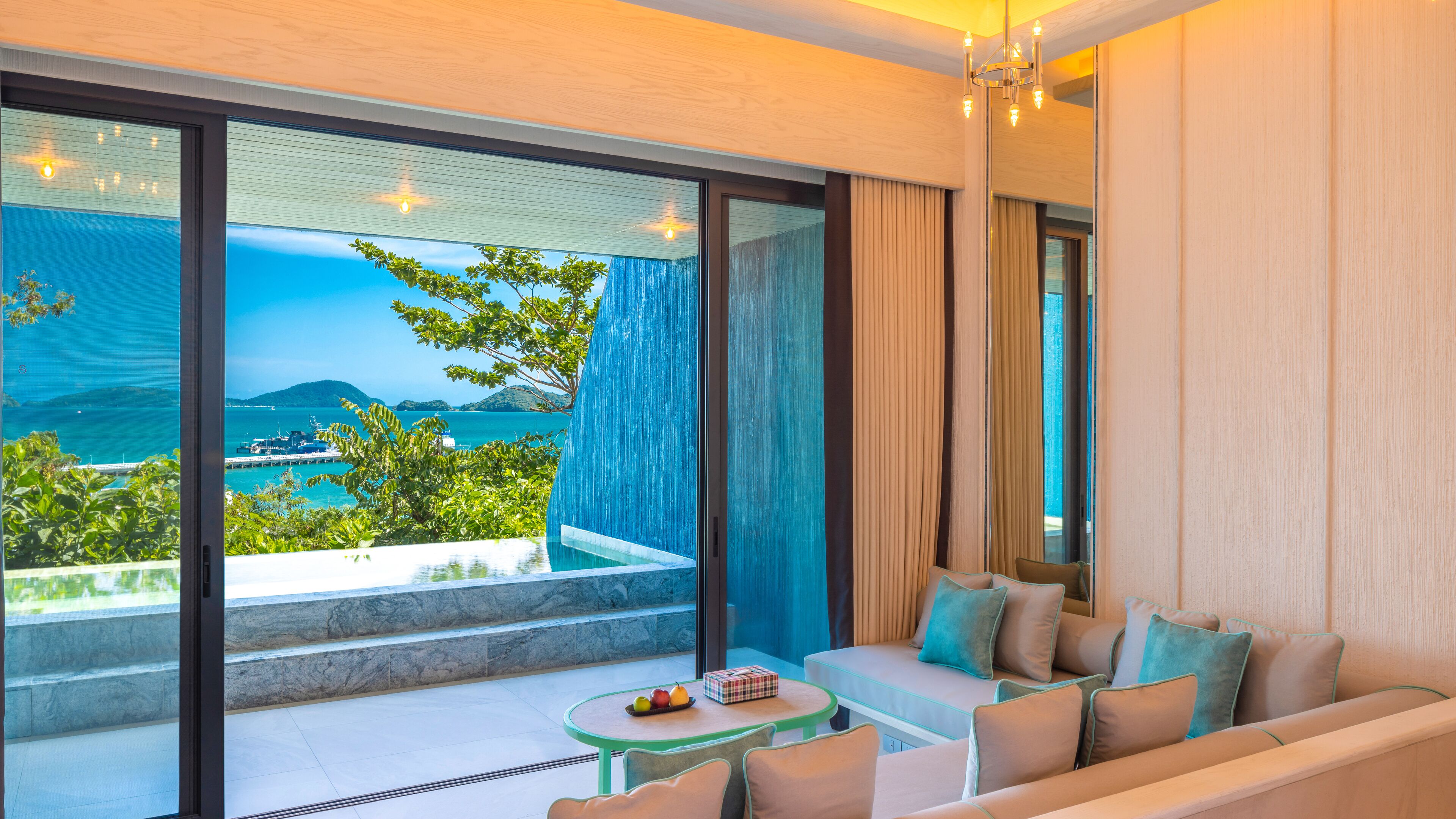 Habitación de hotel moderna con balcón, sofá, una mesa con frutas y vistas al mar y a las montañas.