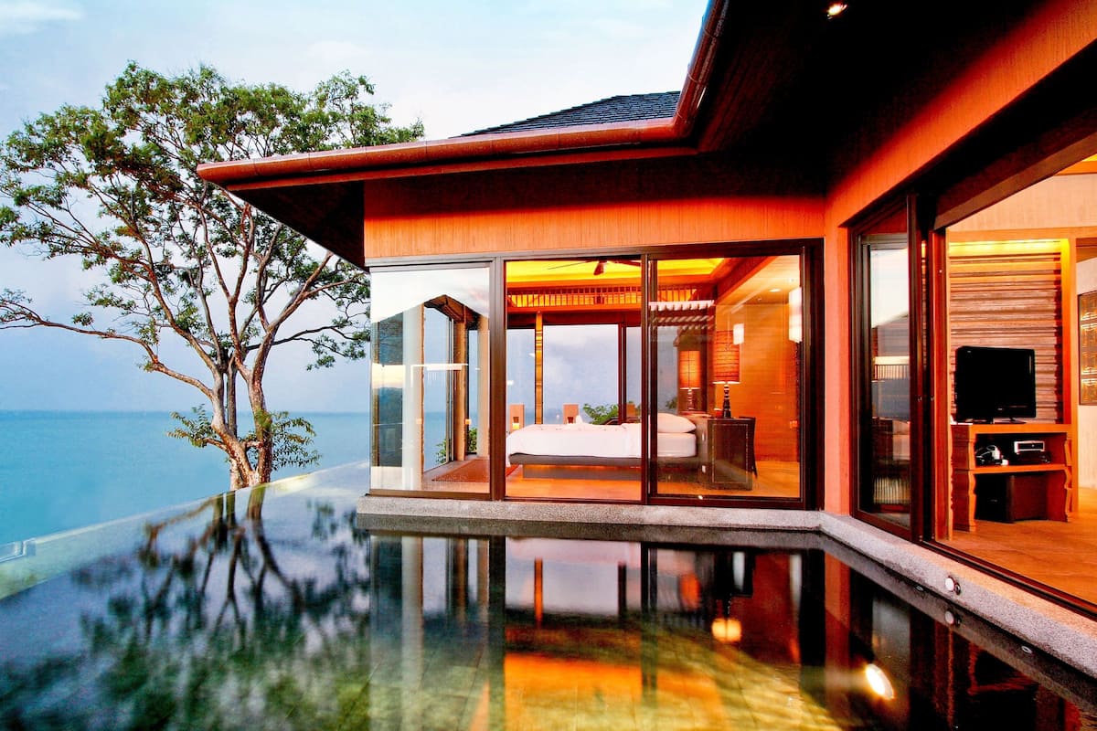 A modern hotel with a glass infinity pool, a bedroom with a large bed and a TV, and a view of the sea.