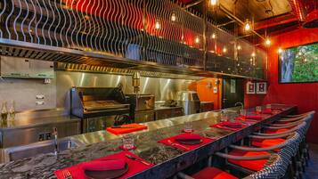 A restaurant with a long counter, red chairs, and a modern kitchen in the background.
