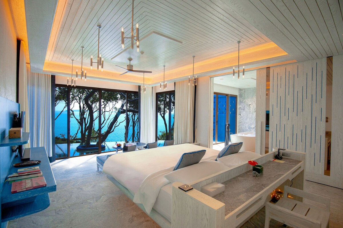 A modern hotel room with a large bed, a desk with a computer, and a view of the sea.