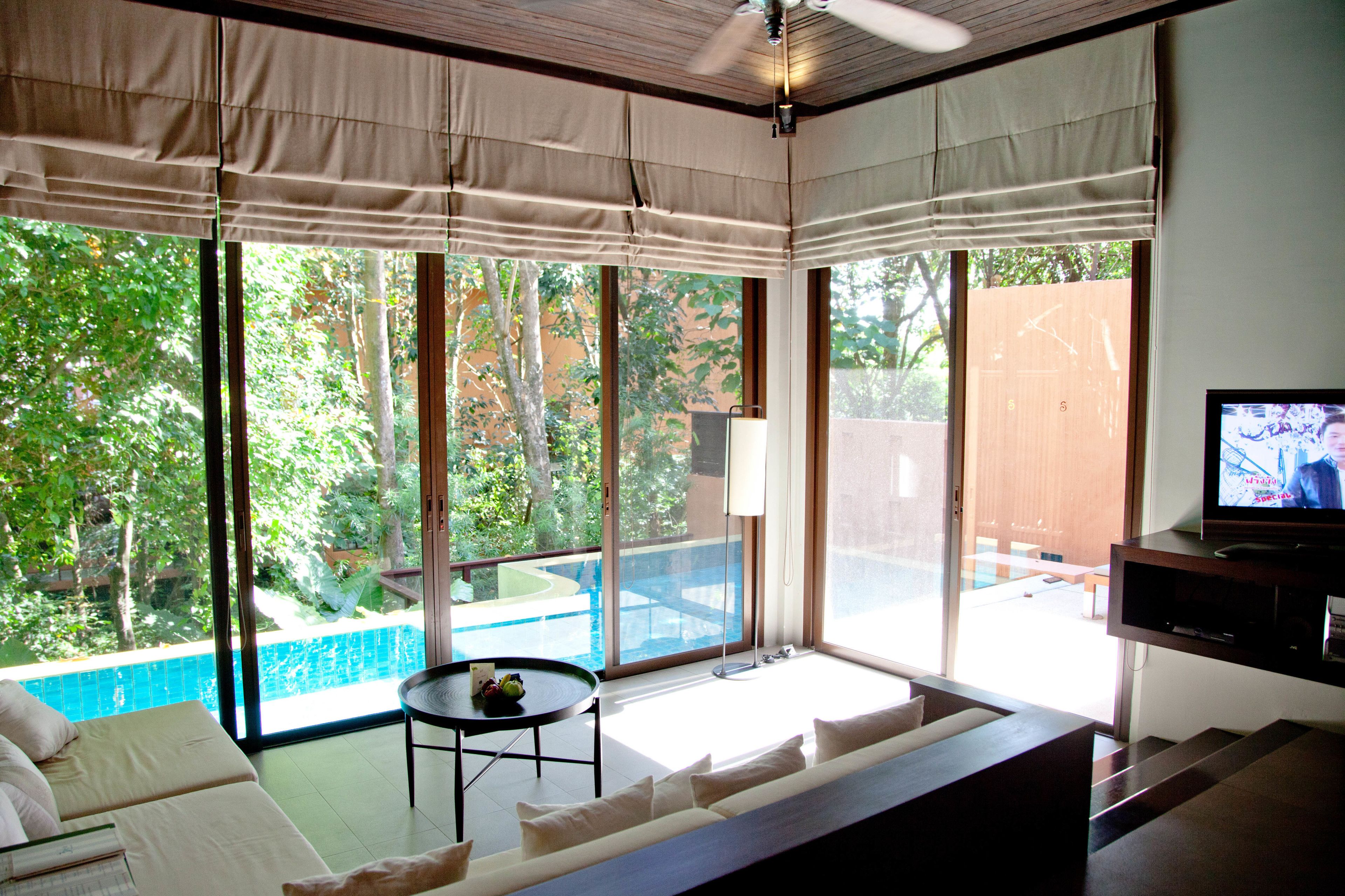 A modern living room with a large sliding glass door leading to a pool area, a sofa, a small table with a bowl of fruit, and a television on a wooden stand.