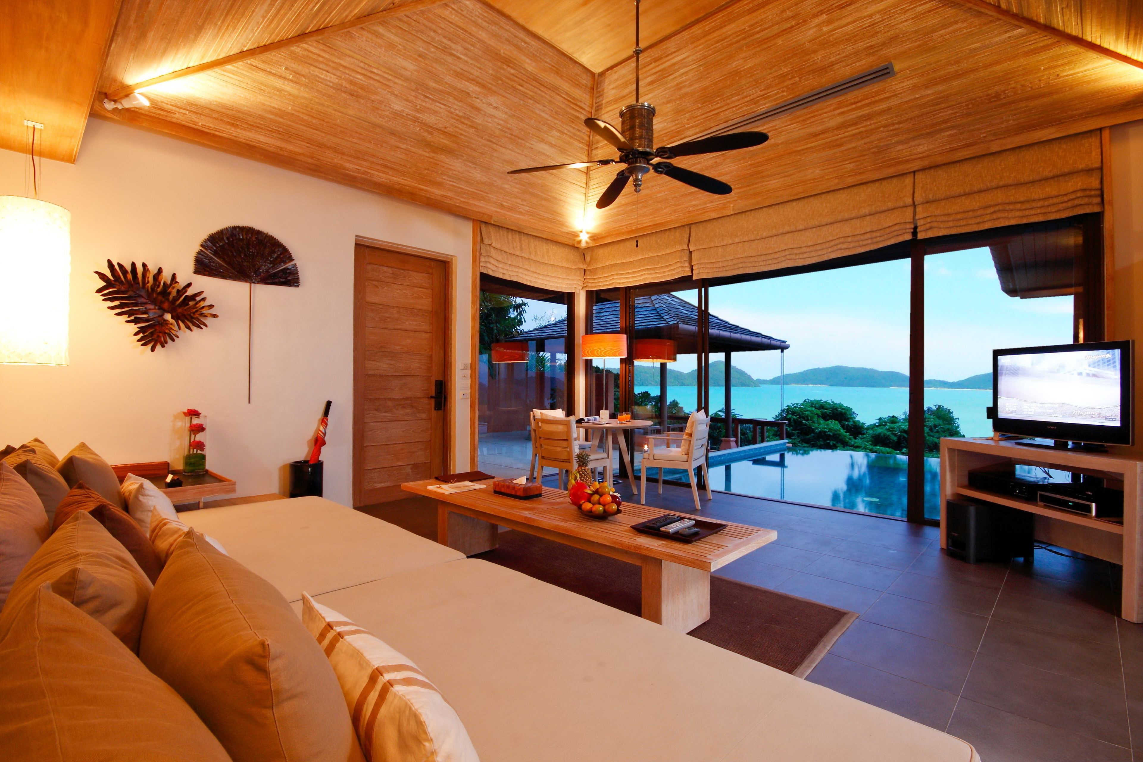 A modern living room with a large sofa, a dining area, and a TV. There is a ceiling fan, a large window with a view of the ocean, and a wooden ceiling.