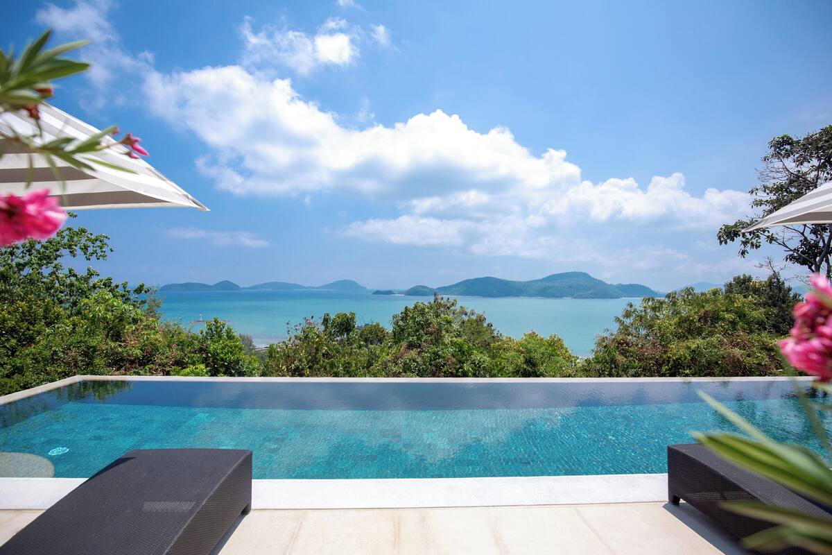 A clear blue sky, a swimming pool with lounge chairs, and a view of the sea and mountains.
