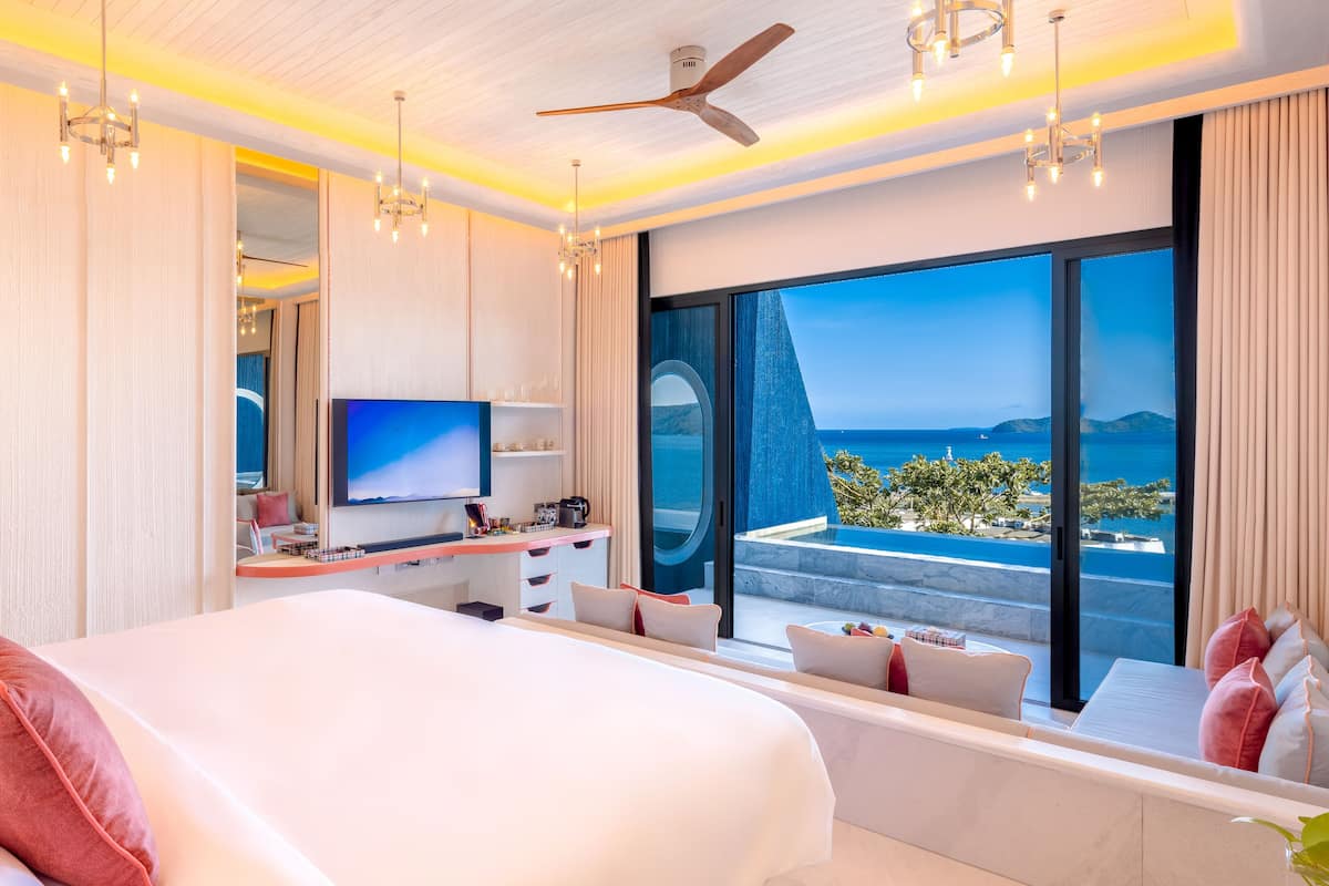A modern hotel room with a large bed, a desk with a flat-screen TV, and a balcony with a sea view.