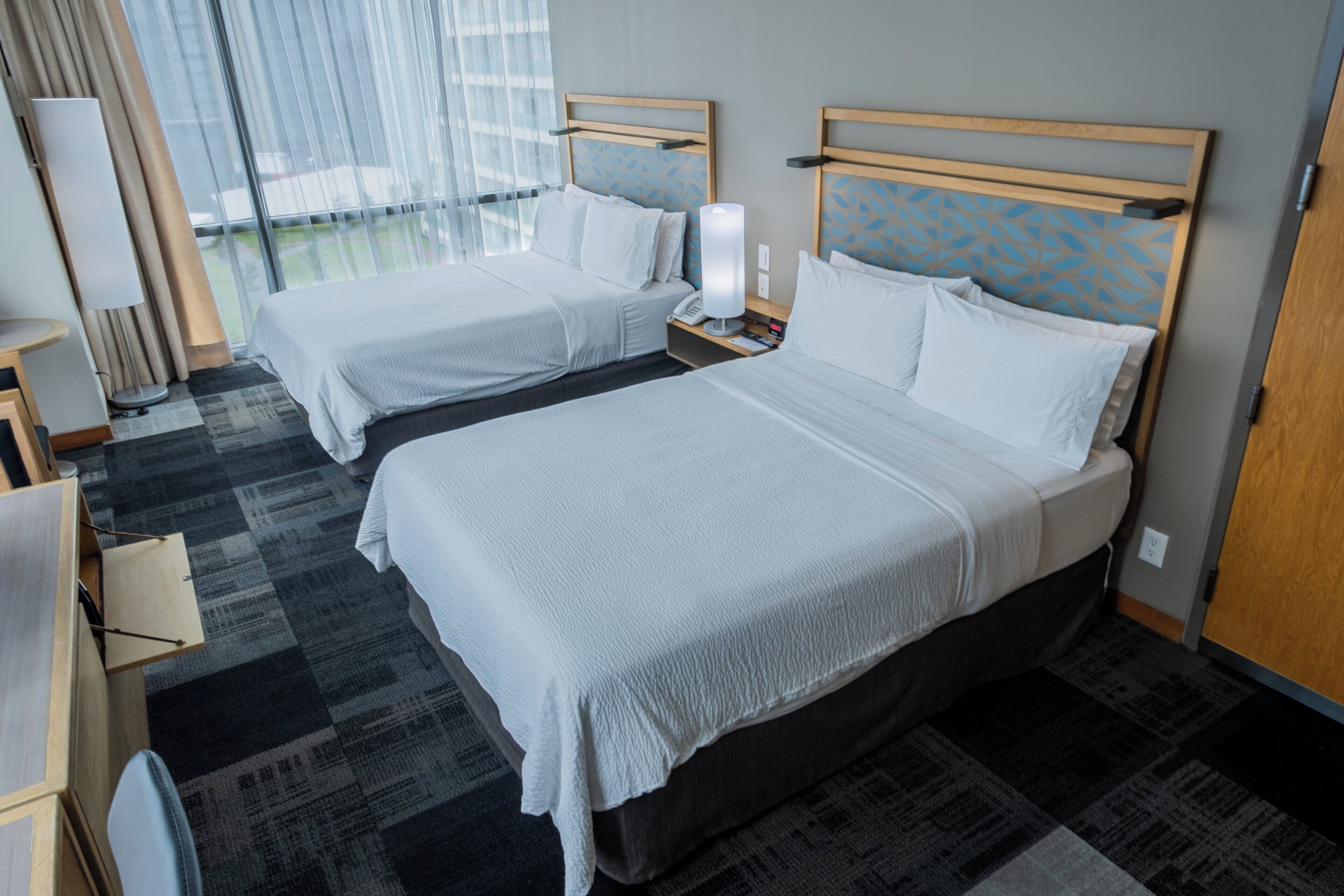 Habitación de hotel con dos camas, una mesita de noche con lámpara y vistas al exterior a través de amplias ventanas.