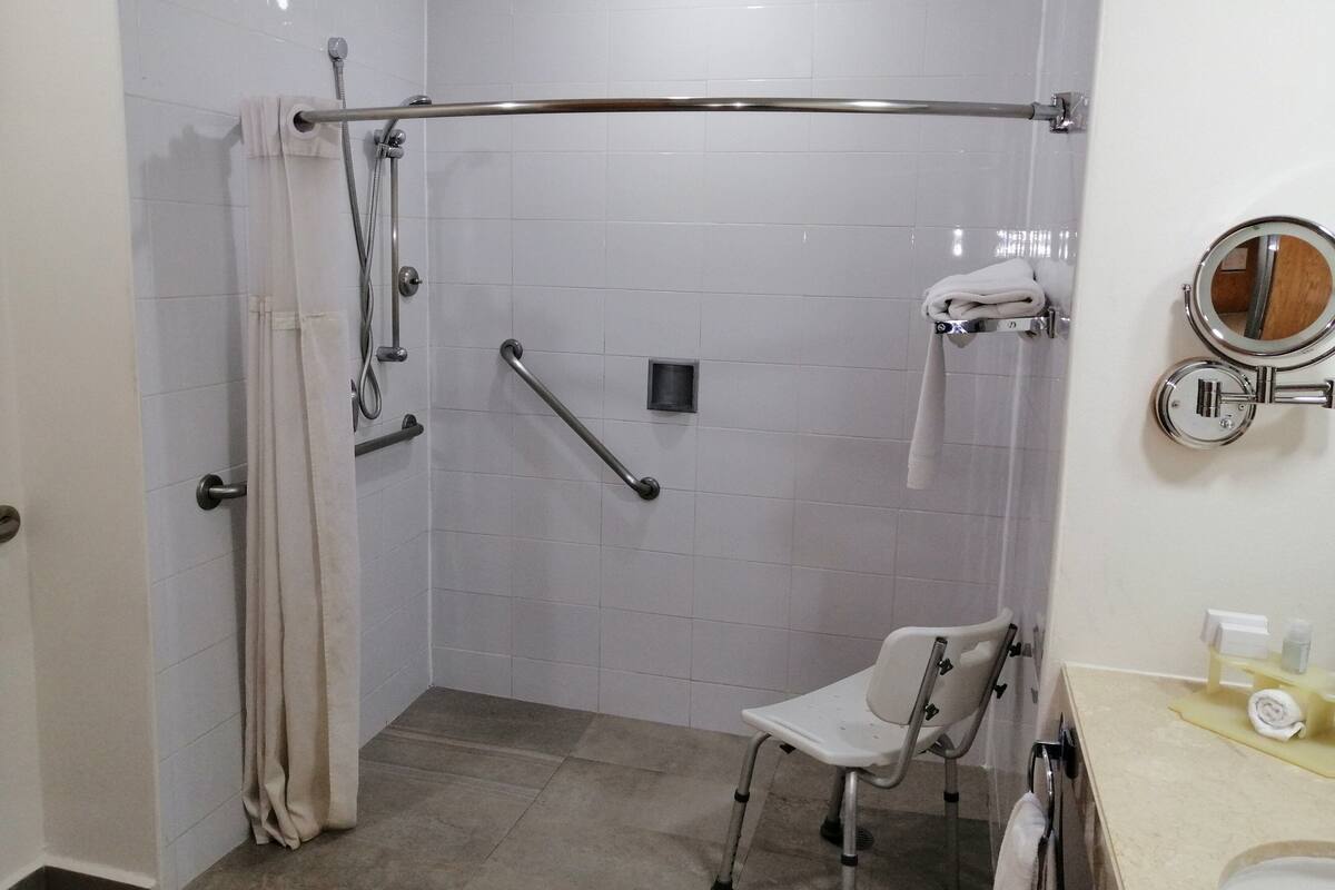 A bathroom with white tiled walls, a shower with grab bars, a wheelchair-accessible chair, and a sink with a mirror.