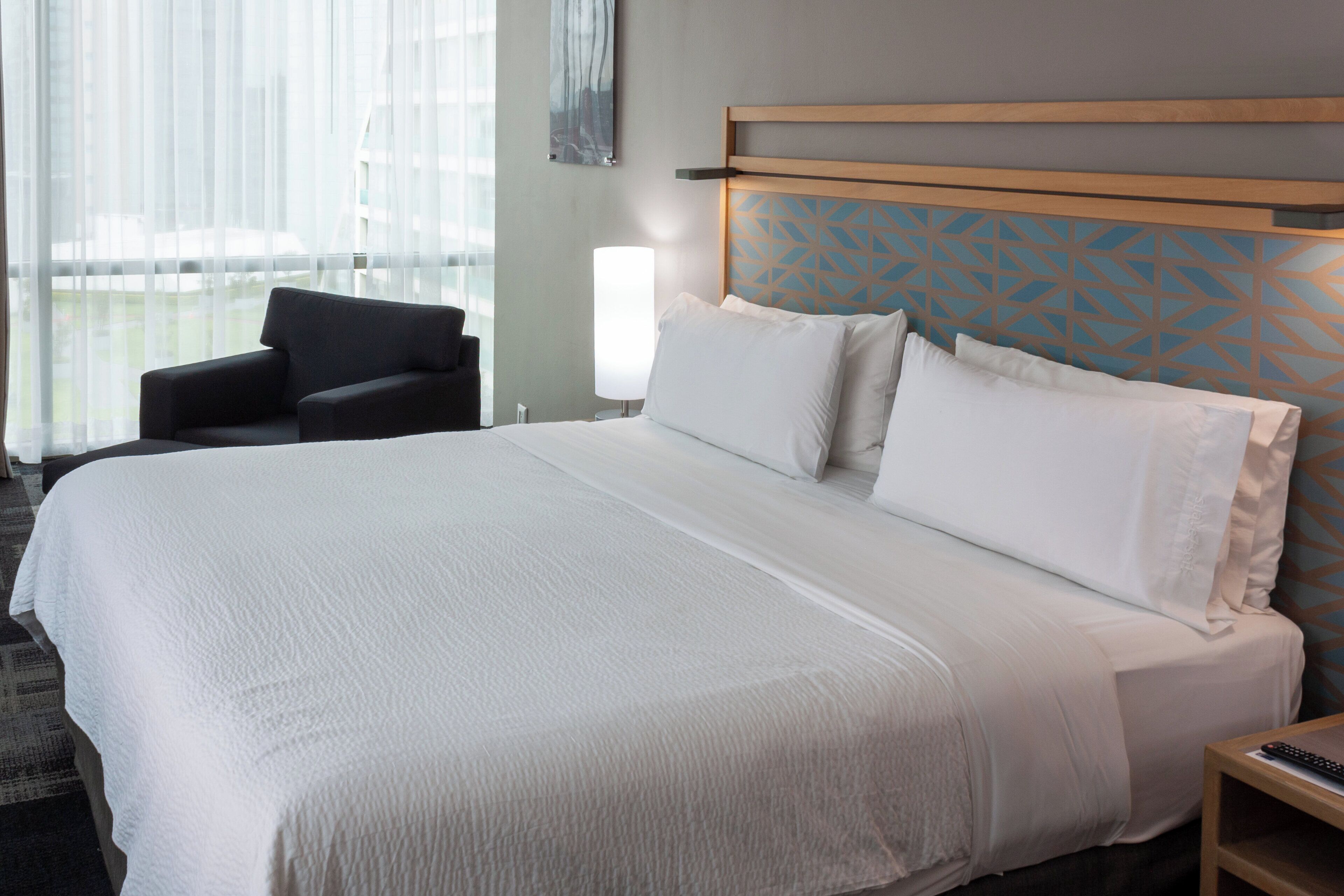 A hotel room with a large bed, a bedside table, a chair, and a window with curtains.