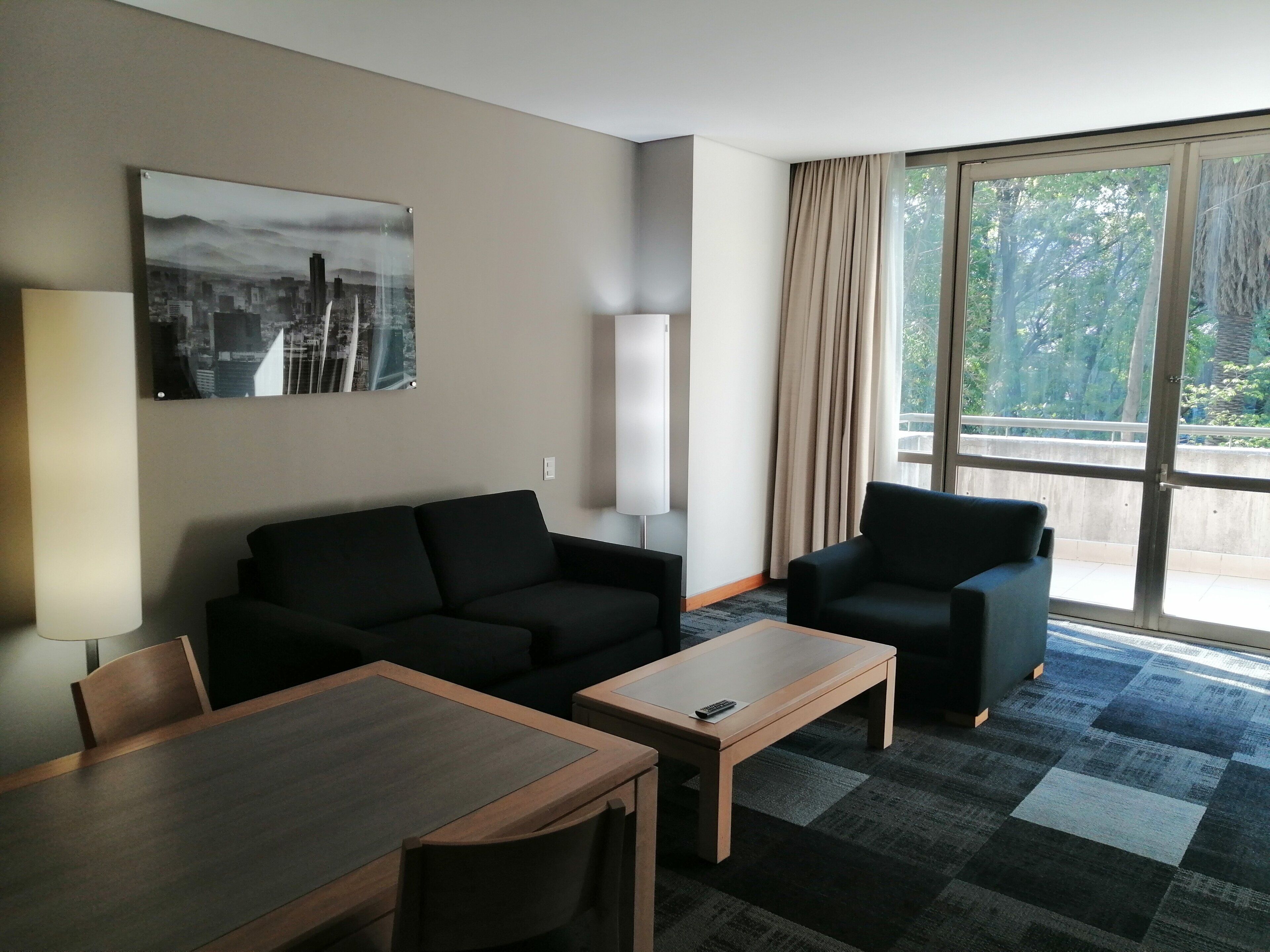 A hotel room with a sofa, a chair, a small table, and a large window with a view of greenery.