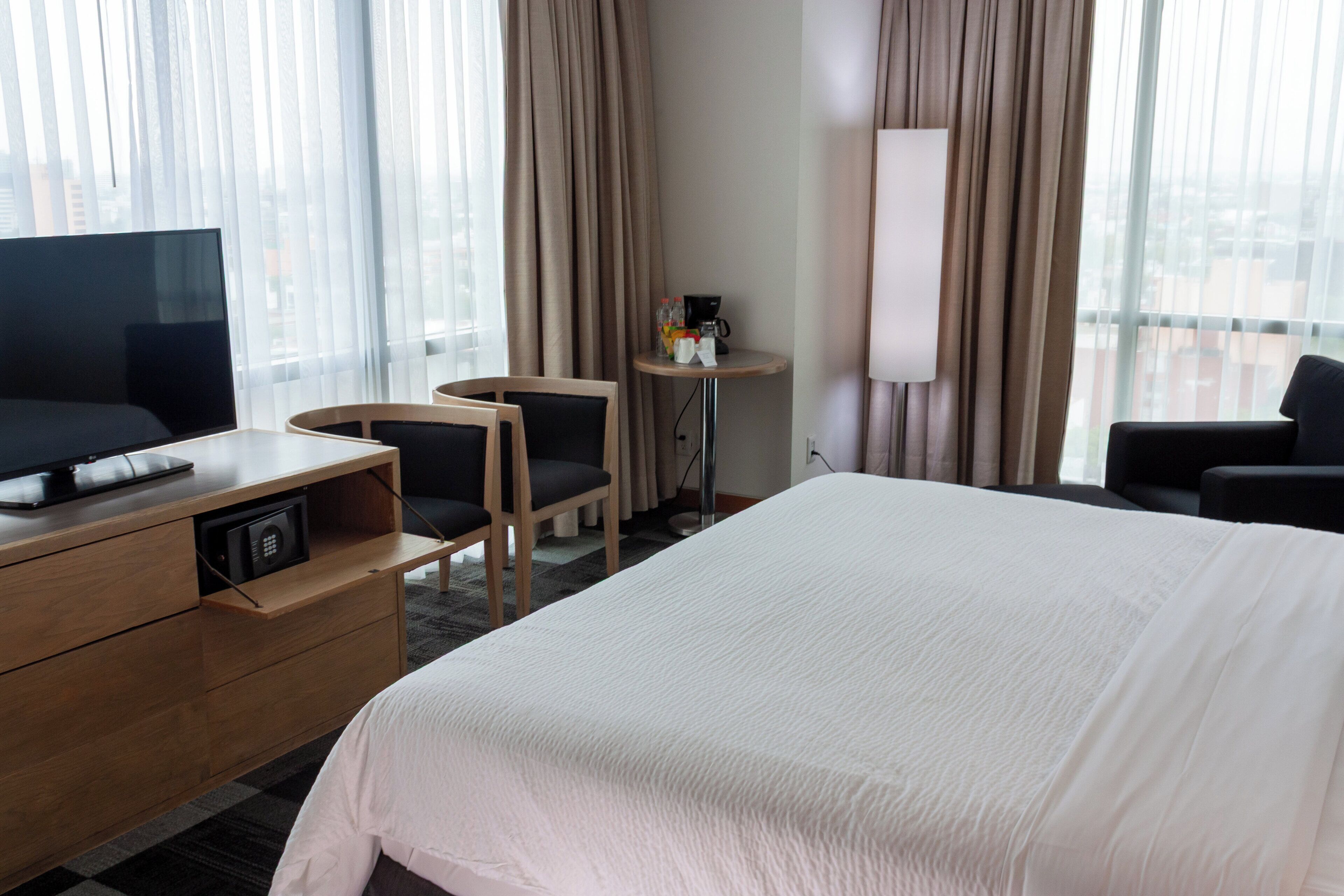 A hotel room with a large bed, a TV on a wooden dresser, two chairs, a small table with a coffee maker, and a lamp.