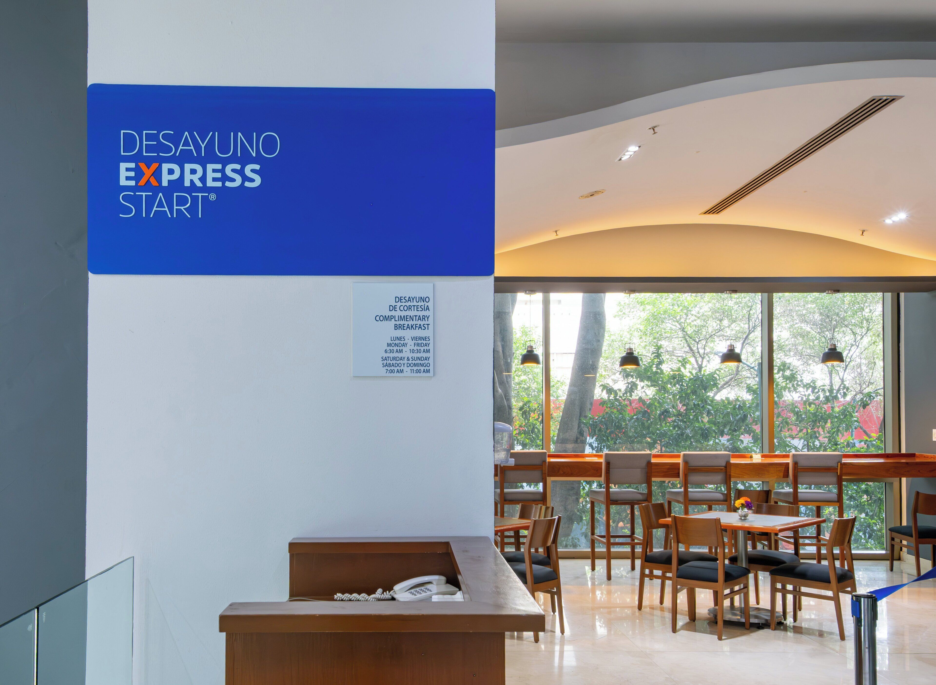 A reception area with a sign for complimentary breakfast, a reception desk, and a seating area with tables and chairs.
