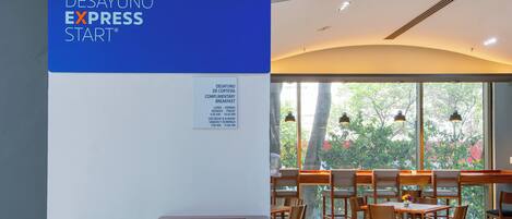 A reception area with a sign for complimentary breakfast, a reception desk, and a seating area with tables and chairs.
