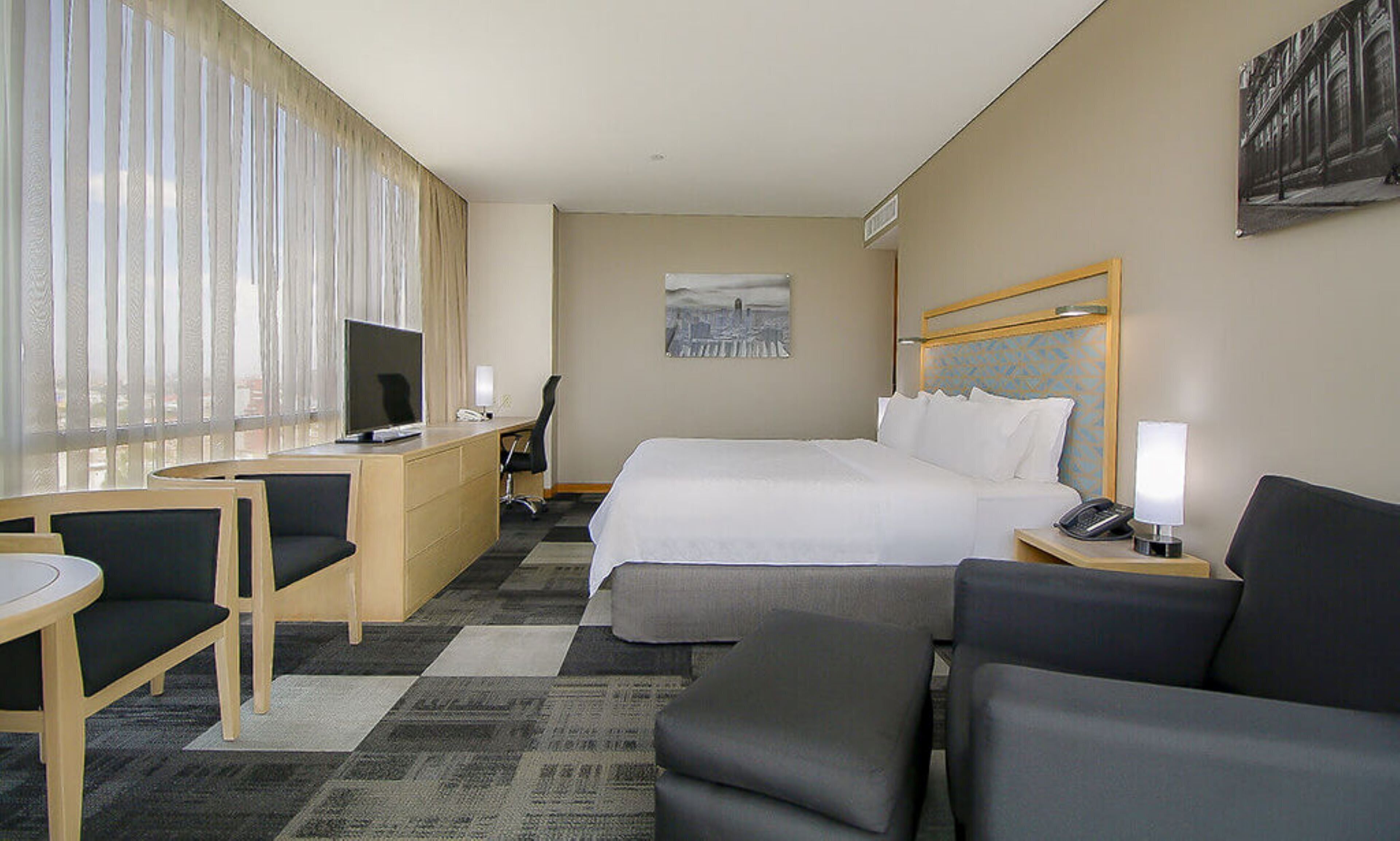 A hotel room with a bed, desk, chair, and a view of the city through the window.