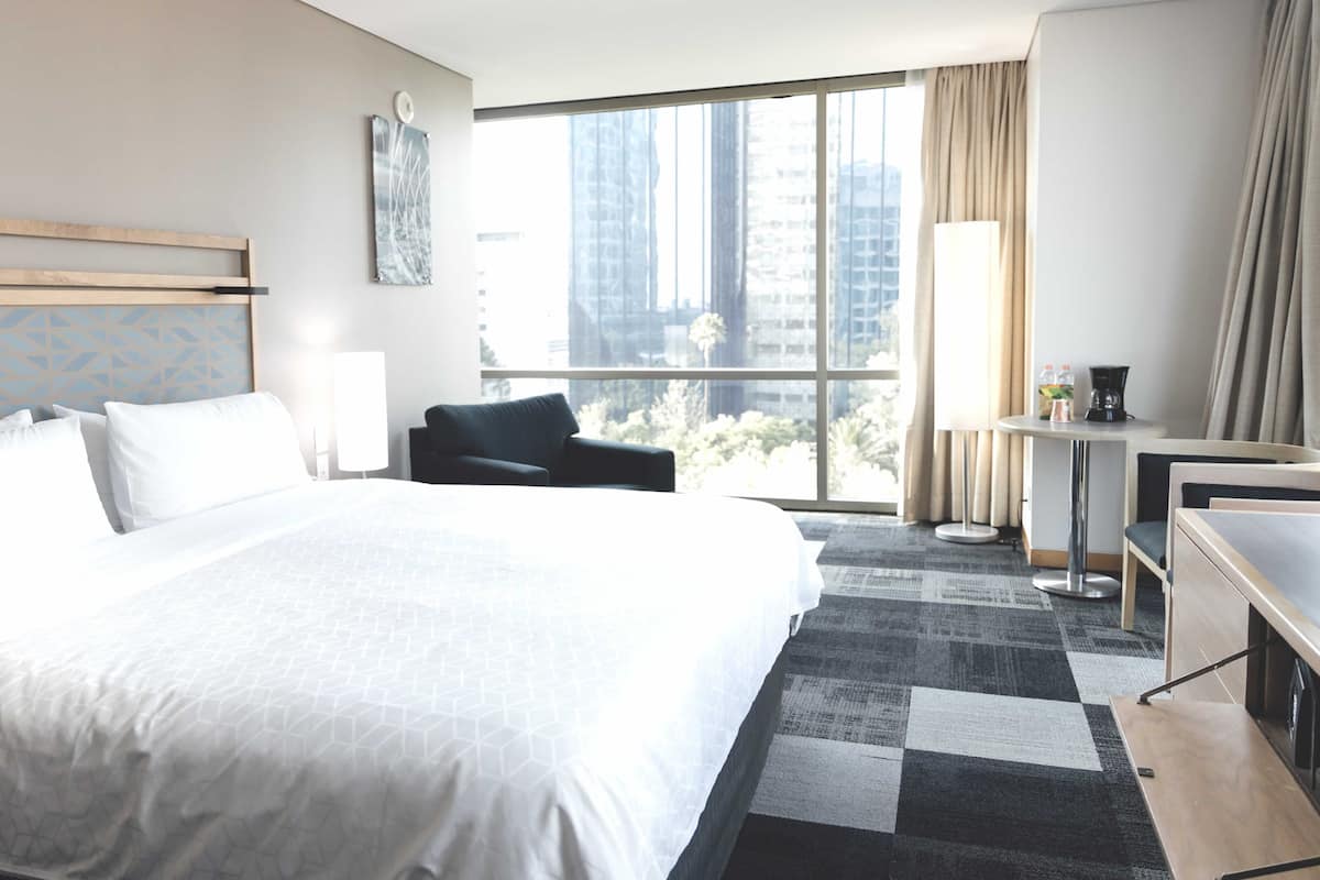 A hotel room with a large bed, a desk, a chair, and a view of tall buildings.