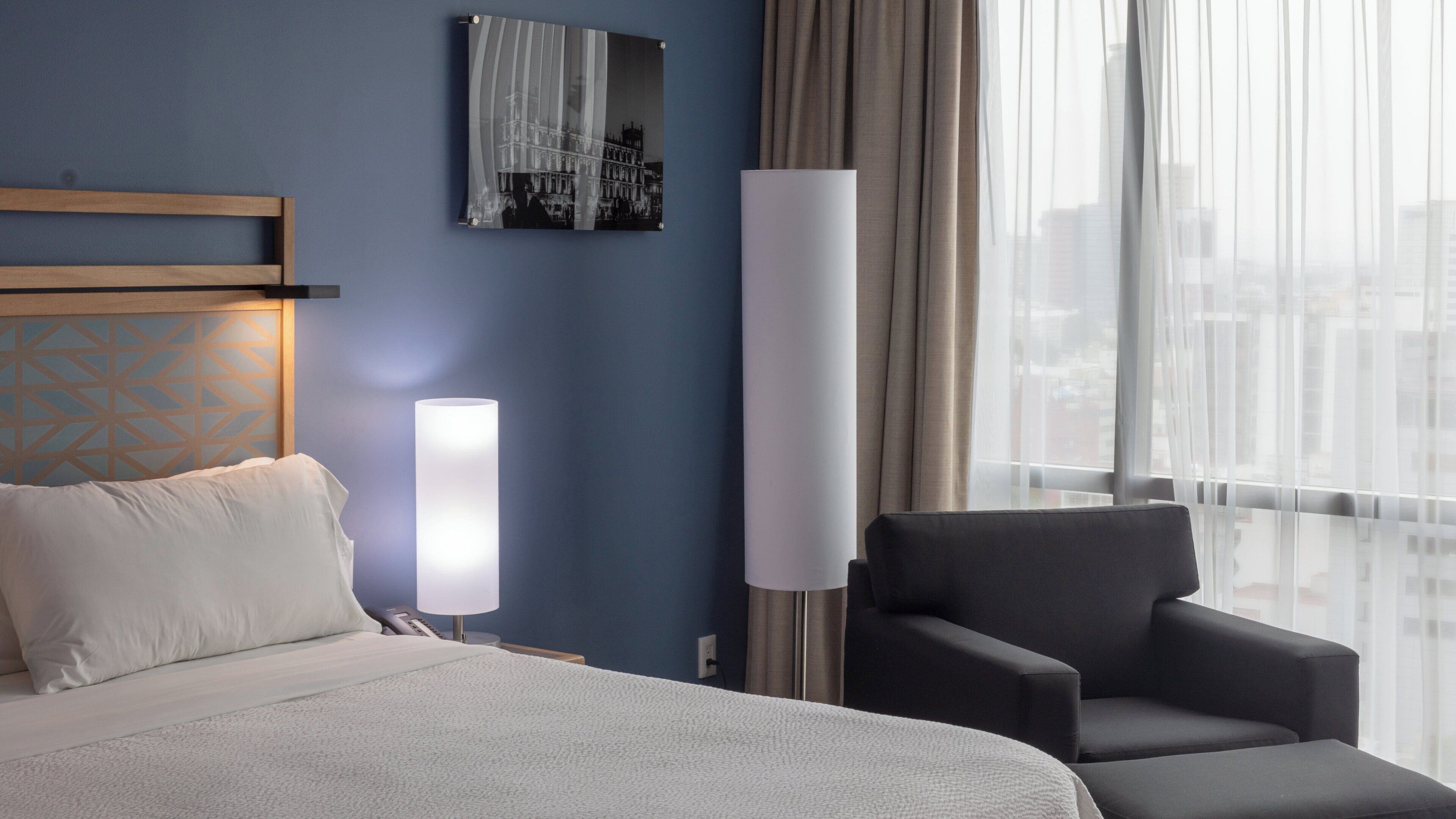 A hotel room with a bed, a chair, a bedside table with a lamp, a wall-mounted picture, and a window with curtains.