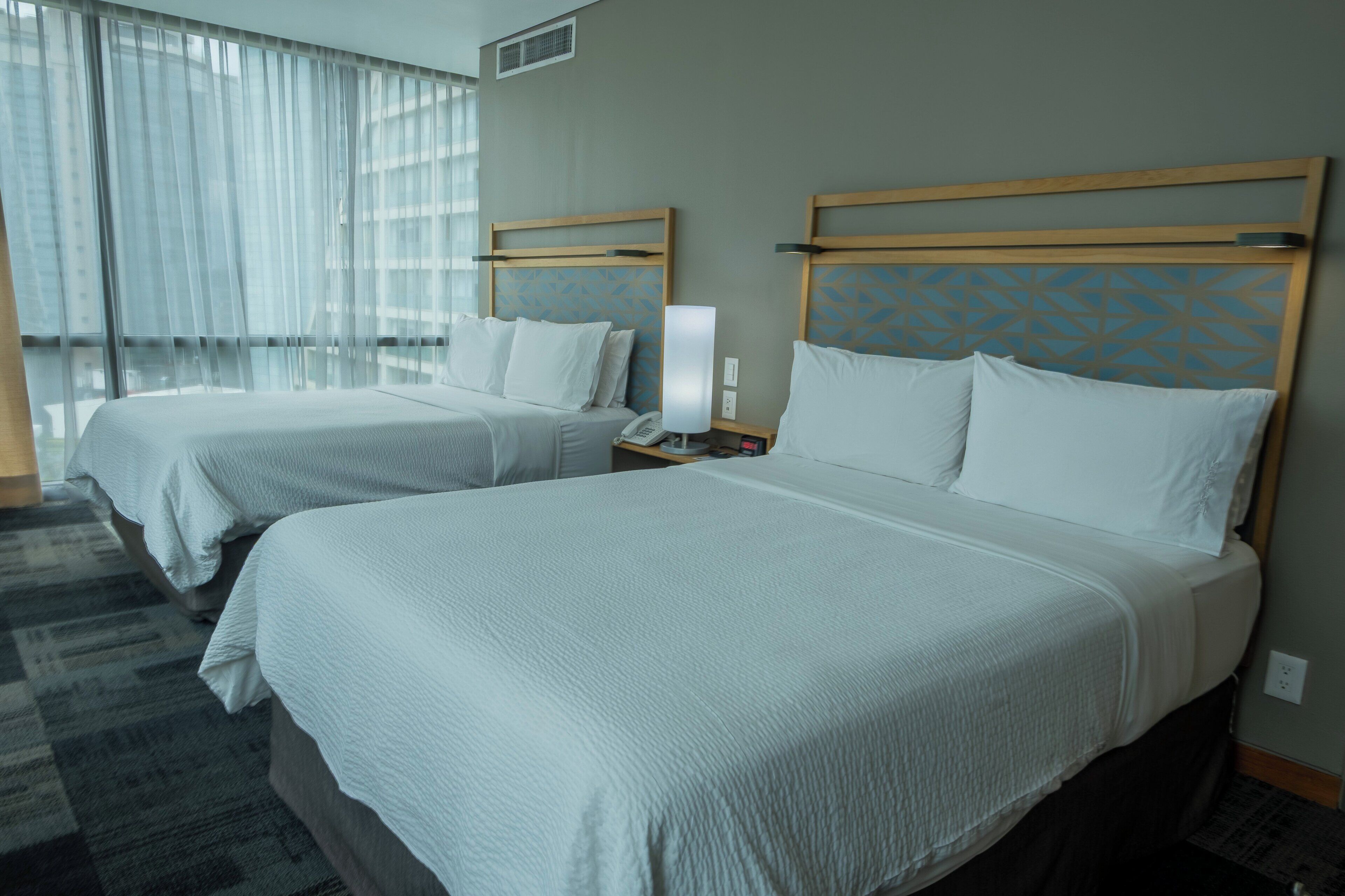 A hotel room with two beds, a nightstand, a lamp, and a window with sheer curtains.