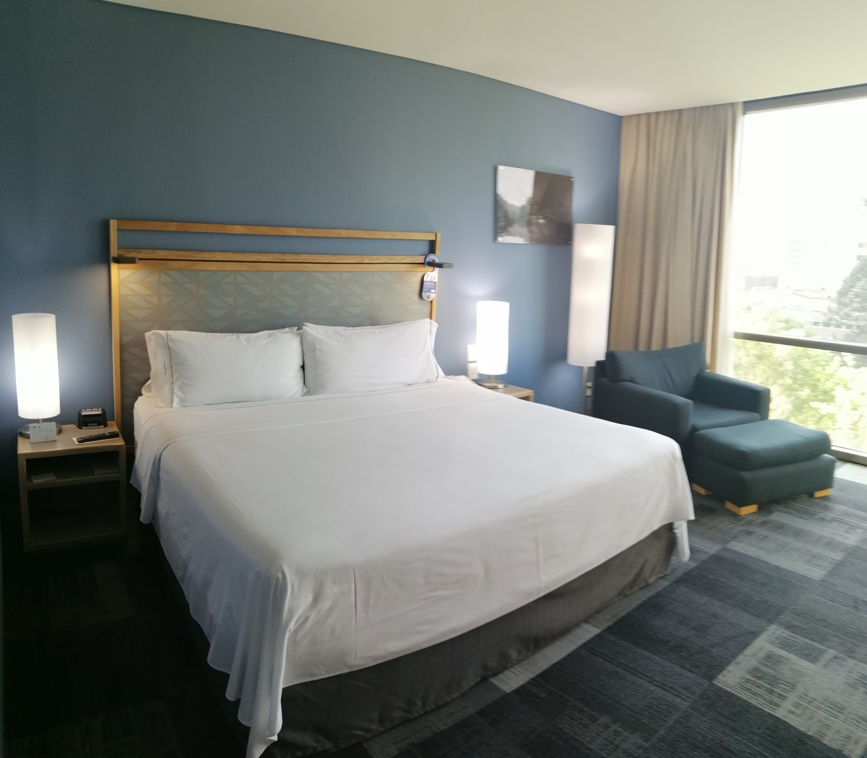 Habitación de hotel con una cama grande, lámparas de noche, una silla y ventana con vistas.