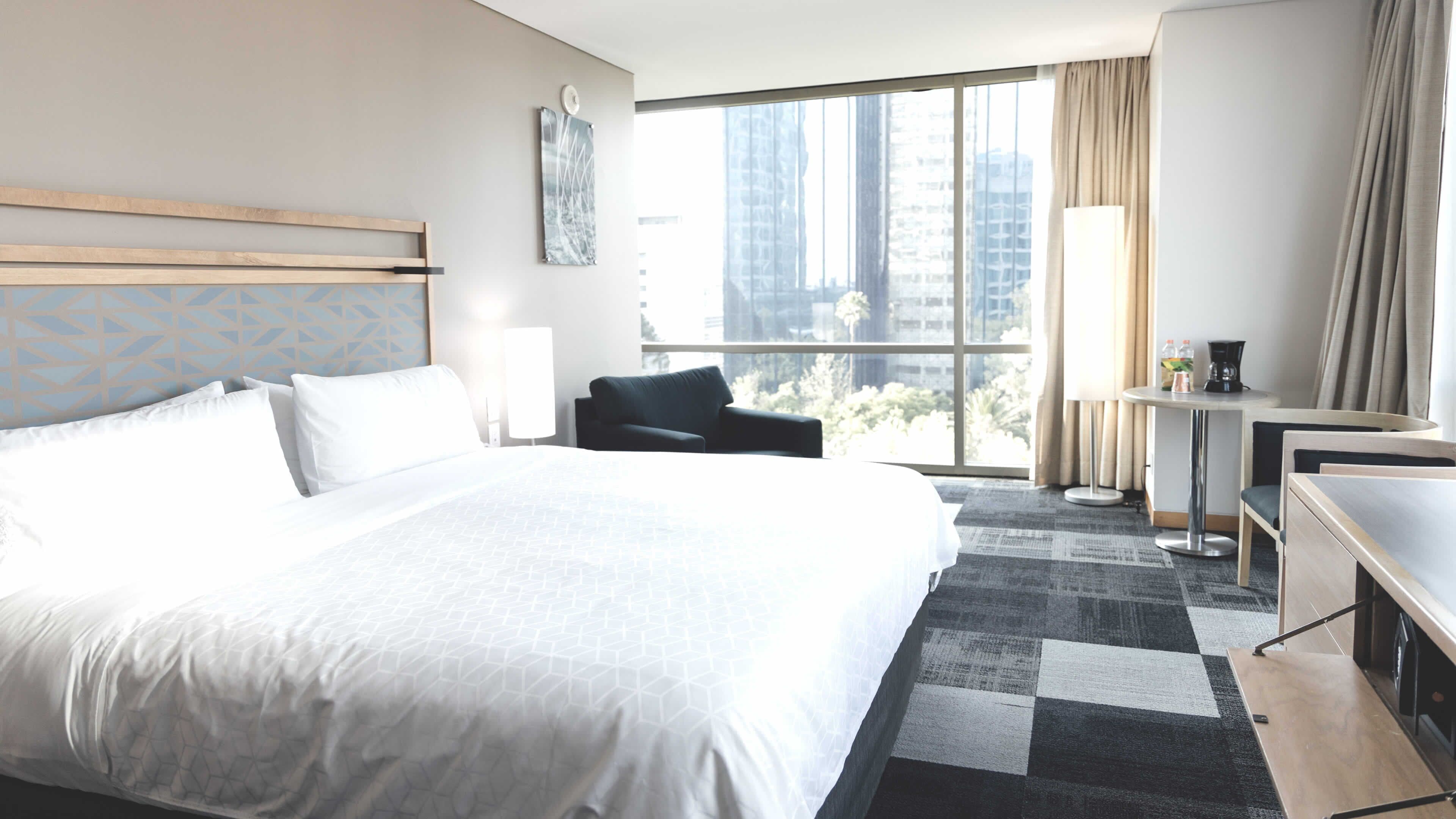 Habitación de hotel con una cama grande, un escritorio, una silla y vistas a la ciudad a través de amplias ventanas.