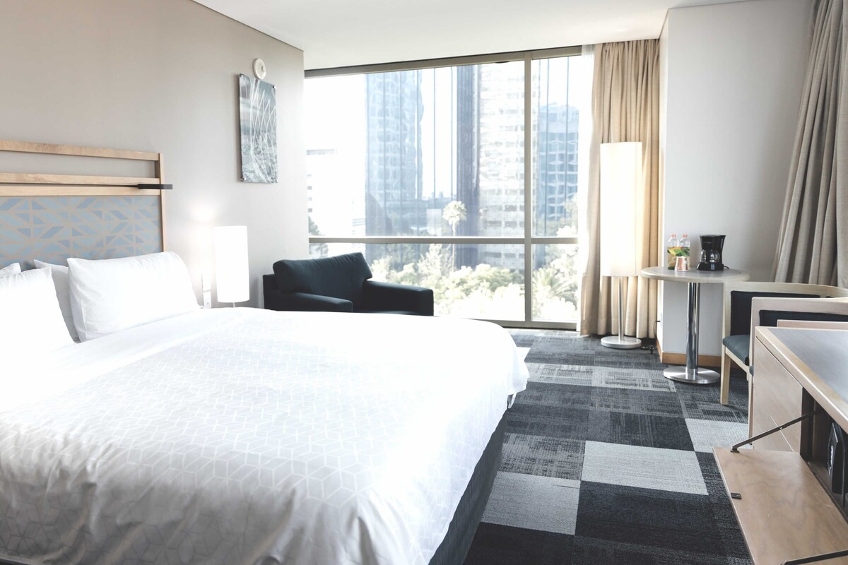 Habitación de hotel con una cama grande, un escritorio, una silla y vistas a la ciudad a través de amplias ventanas.