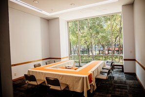 Un salón de conferencias con una larga mesa, sillas y un amplio ventanal con vistas a árboles y edificios.