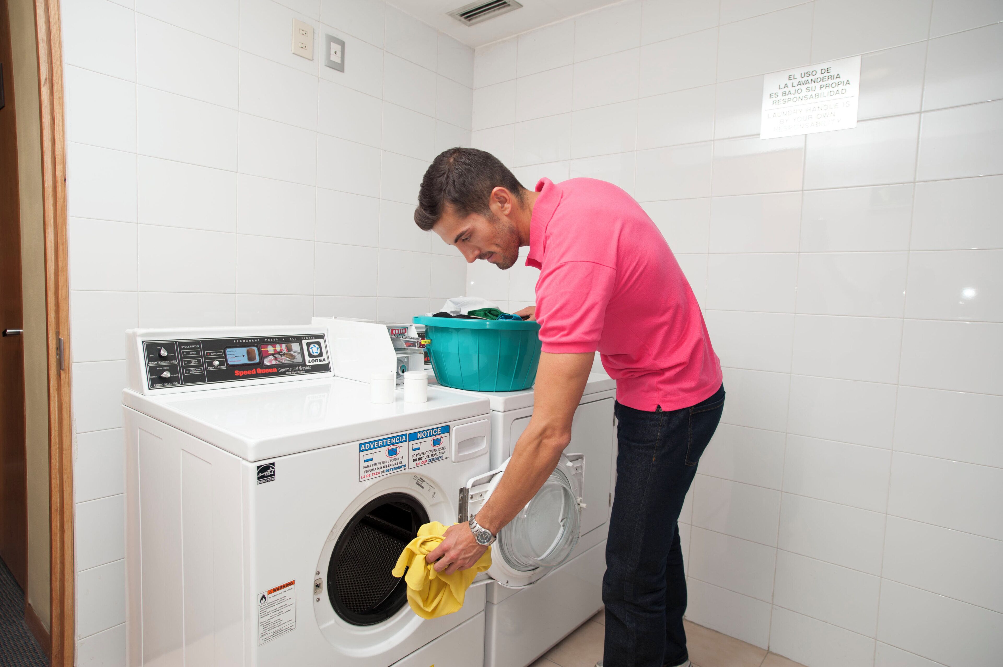 A person loading a yellow garment into a white front-loading washing machine in a room with white tiled walls.