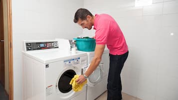 A person loading a yellow garment into a white front-loading washing machine in a room with white tiled walls.