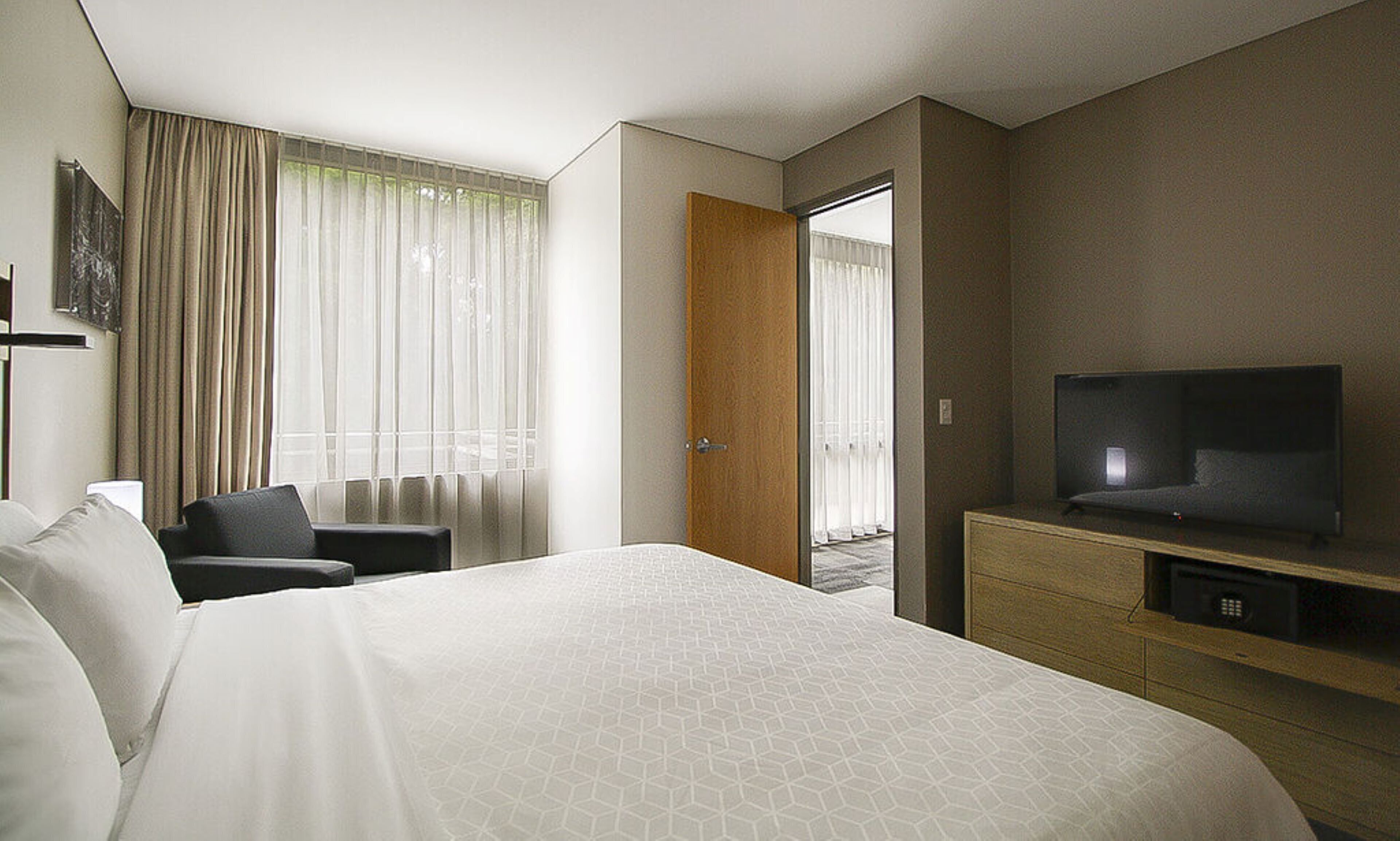 Quarto de hotel com uma cama grande, uma TV sobre um móvel de madeira, uma mesa e uma cadeira.