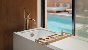 A modern bathroom with a bathtub, a wooden panel wall, and a view of a pool and lounge area.