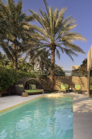 A pool area with a clear blue pool, surrounded by palm trees and a seating area with chairs and a cushioned bench.