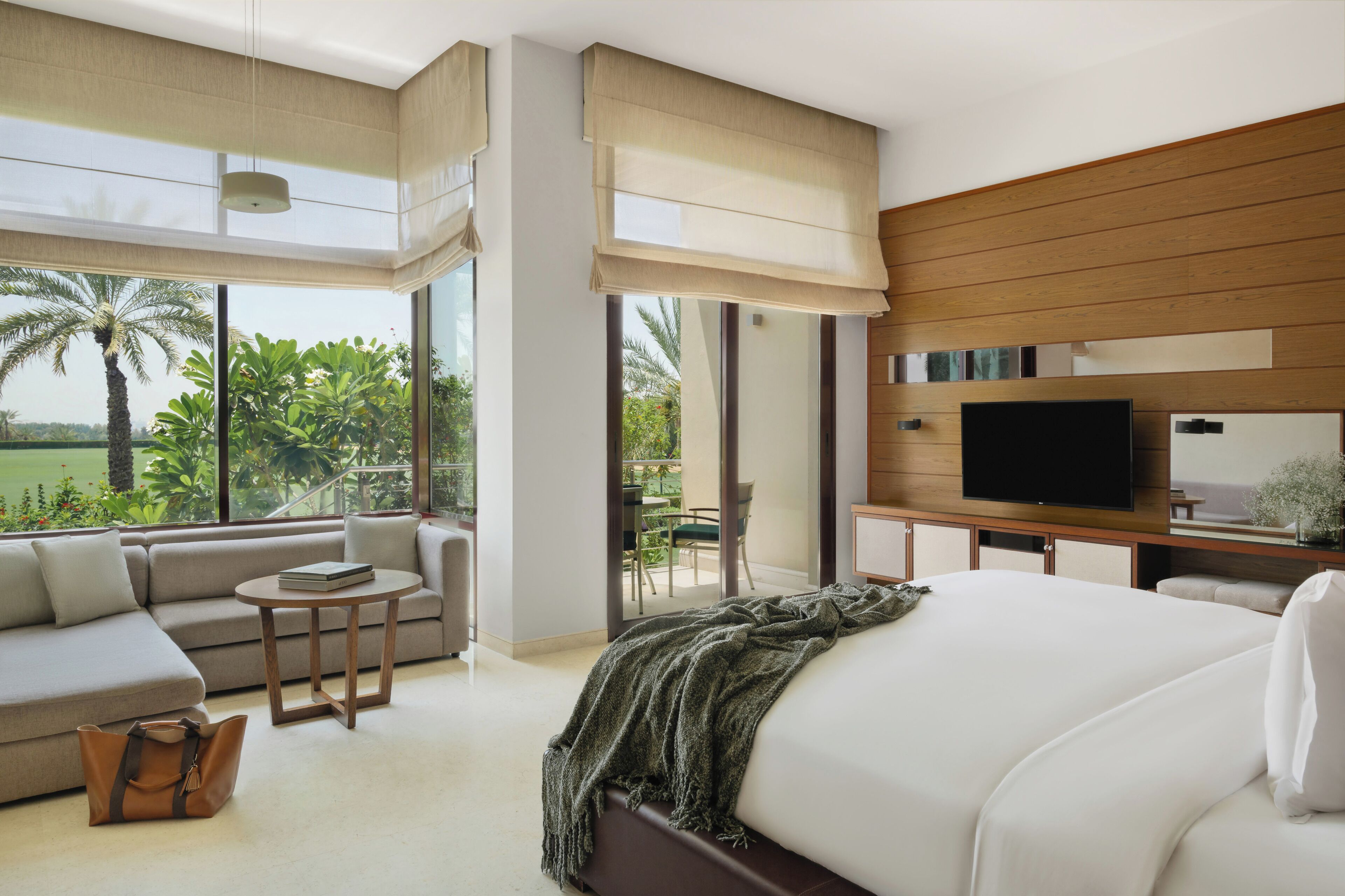 Una habitación de hotel moderna con una cama grande, un sofá, una mesita, un televisor y un balcón con vistas a palmeras.
