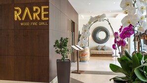 Un intérieur moderne agrémenté d’un panneau indiquant « BARBECUE À FEU DE BOIS RARE », d’une plante en pot et d’un coin salon avec un miroir rond et des coussins.