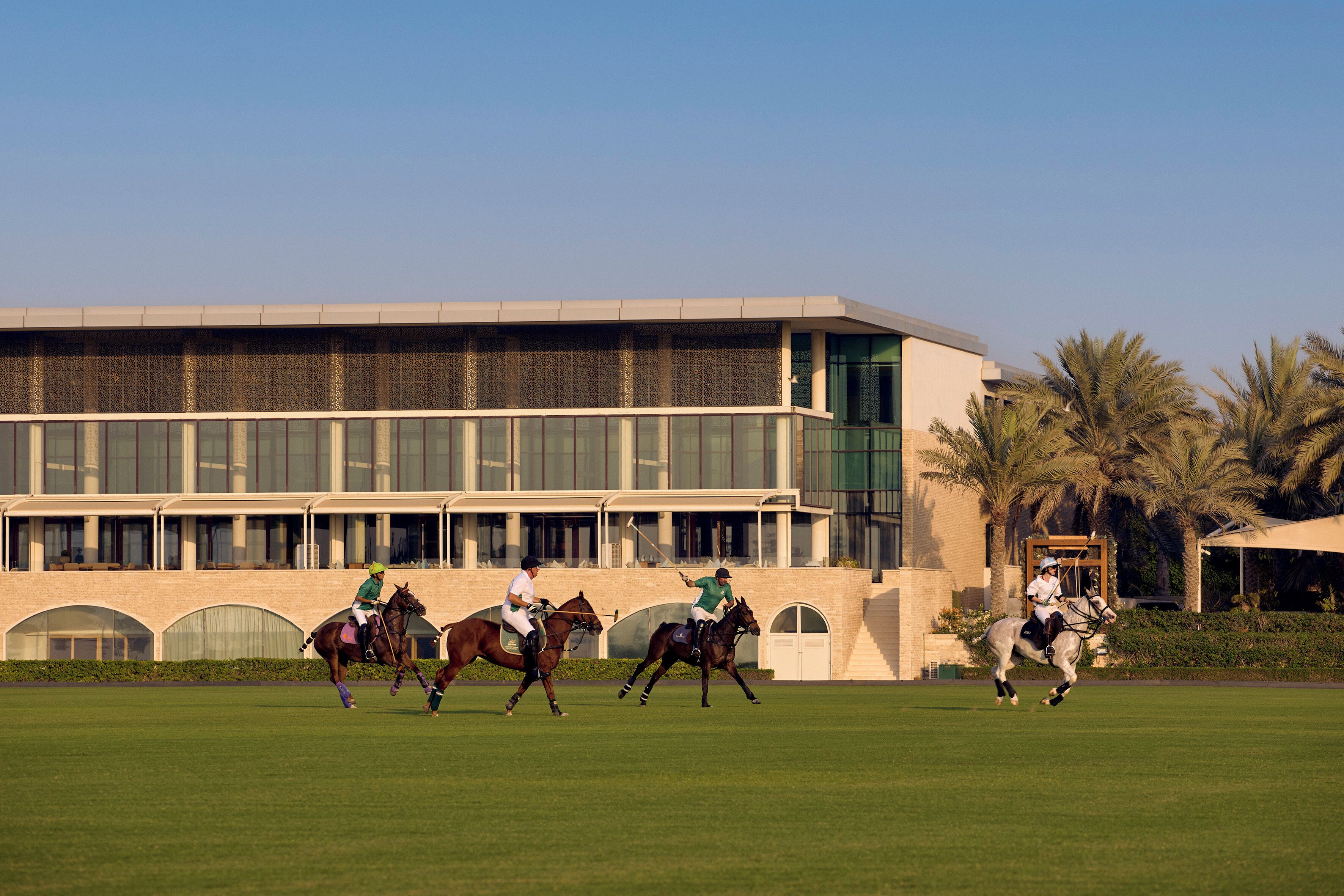 Un campo de polo con jugadores a caballo frente a un edificio moderno con grandes ventanales y una palmera al fondo.