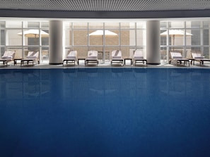 A swimming pool with lounge chairs and umbrellas, large windows, and a brick wall in the background.