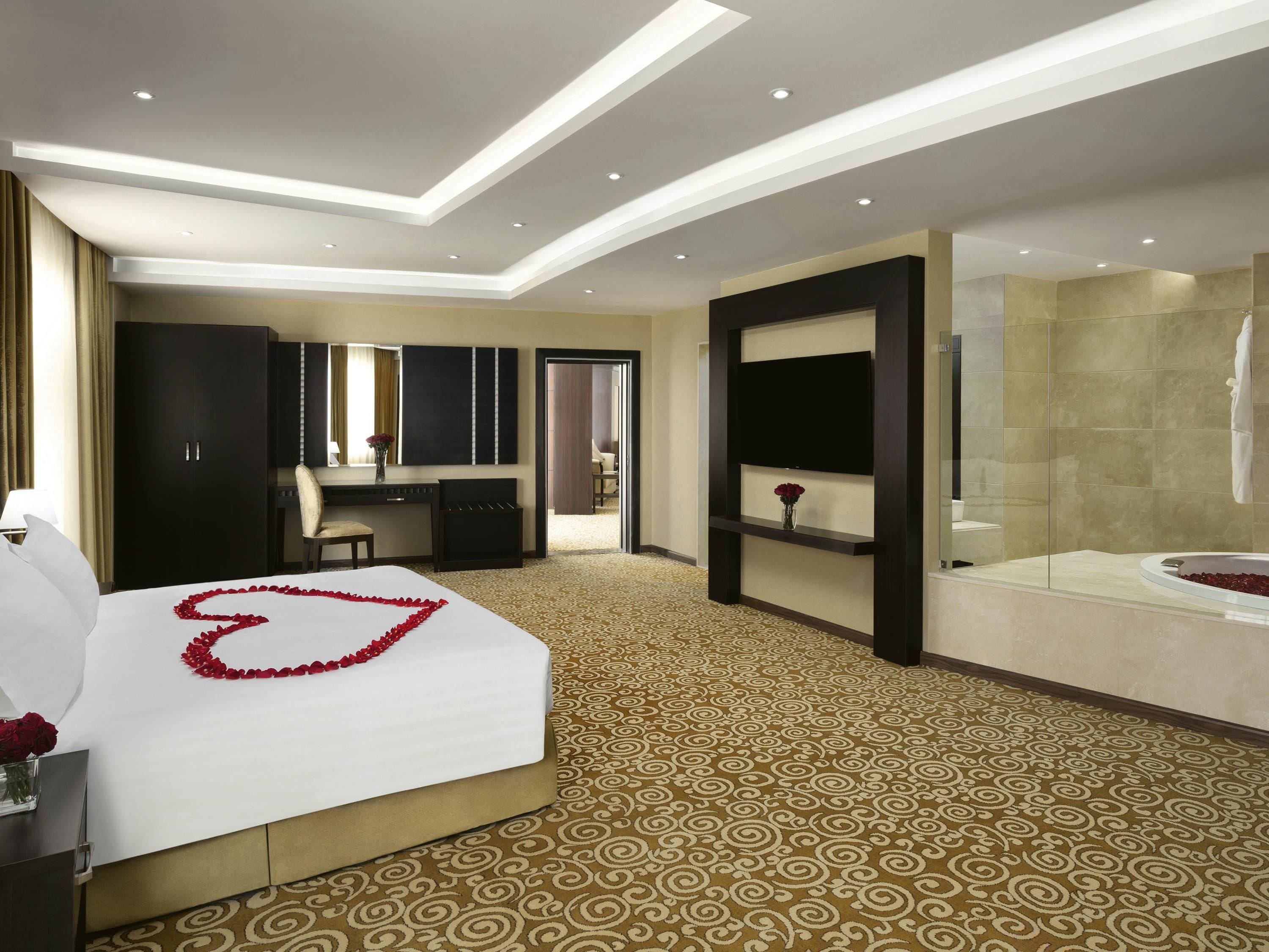 Quarto de hotel com cama, escrivaninha, cadeira, televisão, banheiro e tapete estampado.