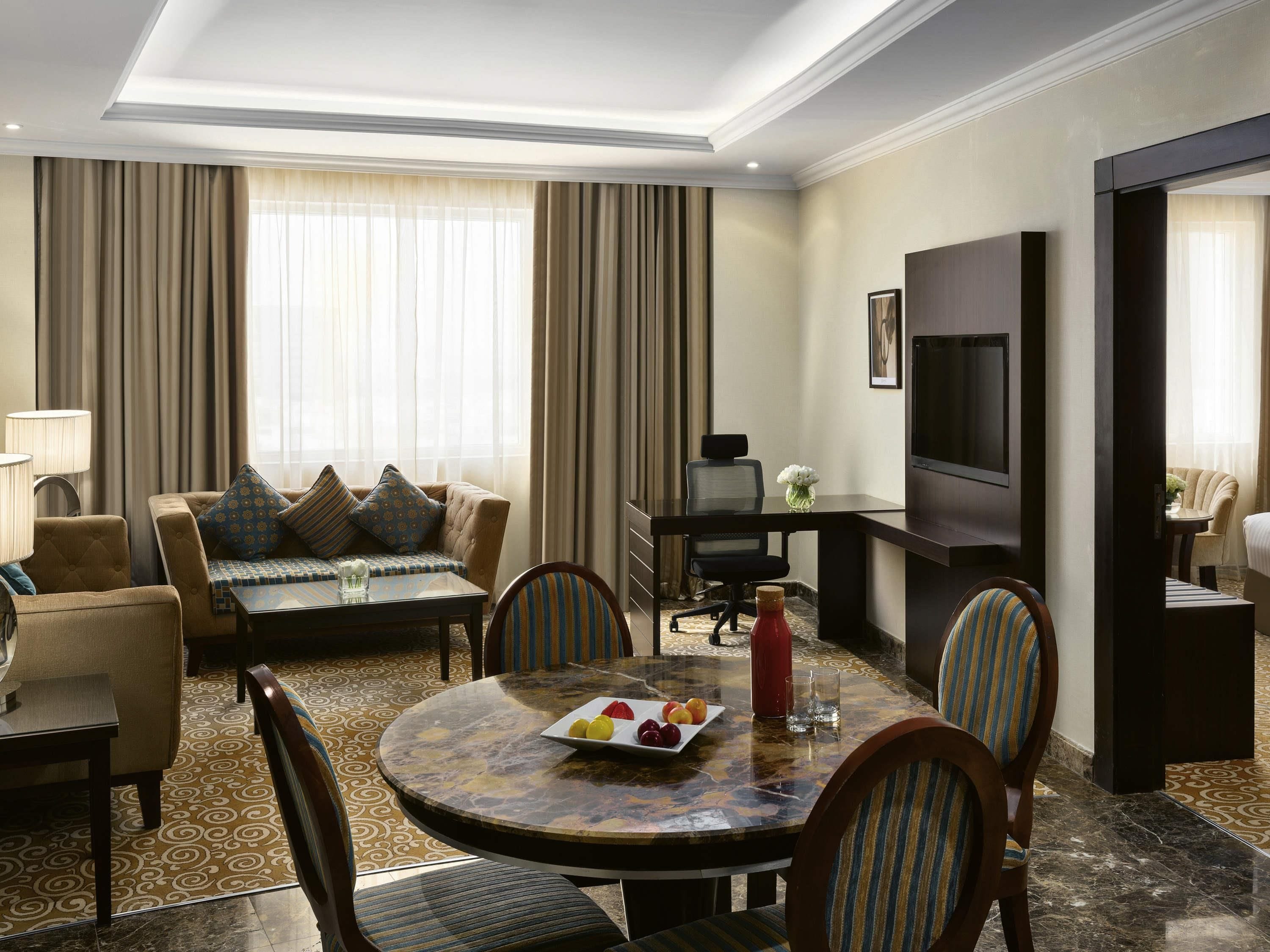 A hotel room with a dining area, a sofa, a TV, and a large window with curtains.
