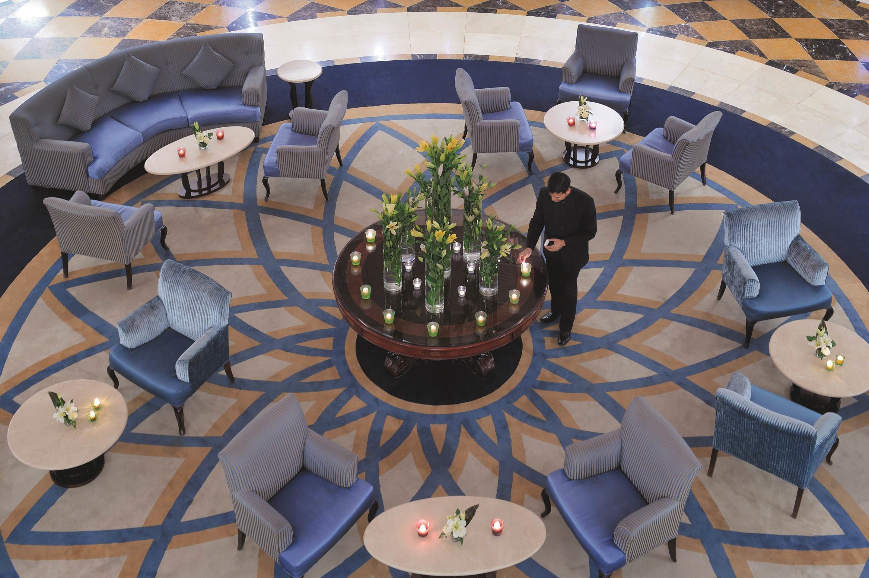 A lobby with a central round table surrounded by sofas and chairs, a patterned floor, and a person standing near the table.
