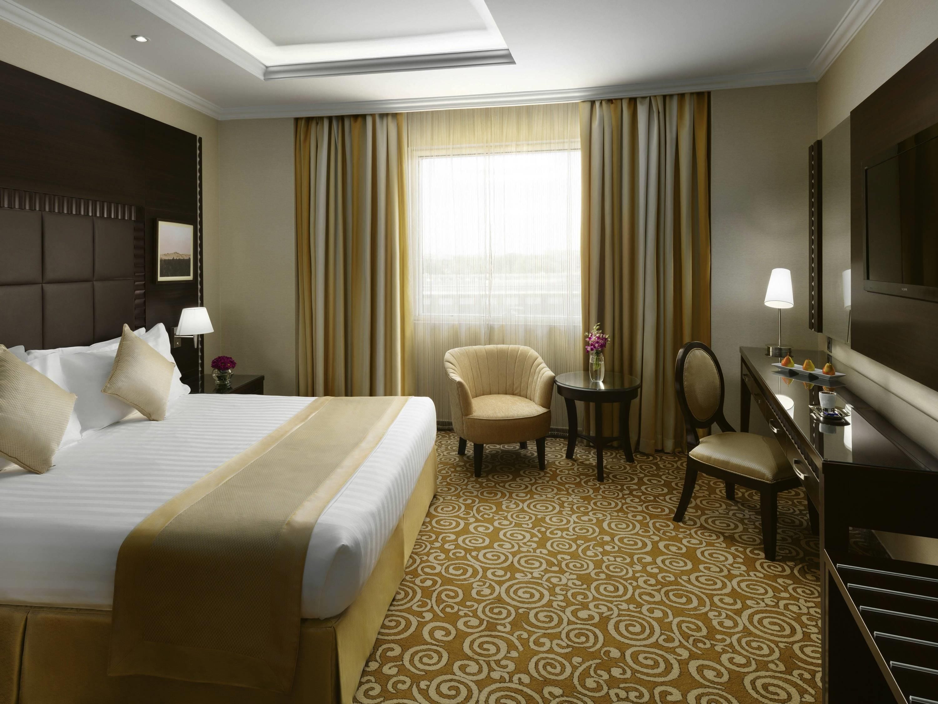 Una habitación de hotel con una cama grande, un escritorio, una silla, una mesita con un florero y un televisor.